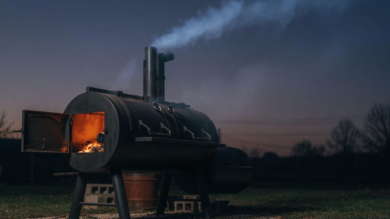 An offset smoker at dusk, firebox glowing, smoke drifting from the stack